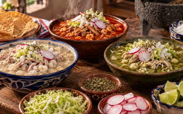 Tres tazones de pozole blanco, rojo y verde con guarniciones al centro de una mesa mexicana.
